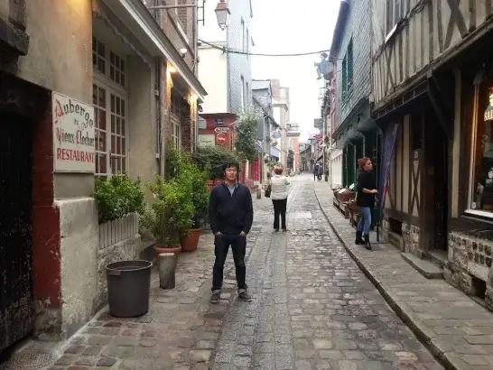 フランス・オンフルール旧市街の石畳の路地に立つ田畑雄貴。歴史的な木組み建築が並ぶ
