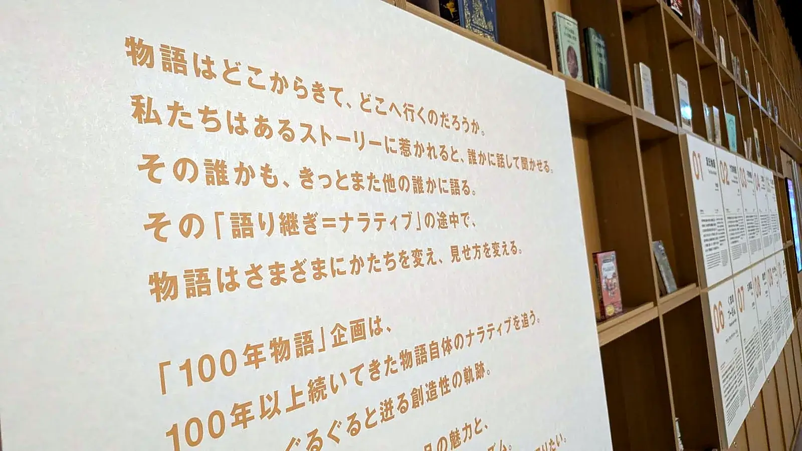MoN Takanawaのライブラリーにある「100年物語」企画の紹介パネルと、物語10作のパネルが並ぶ本棚